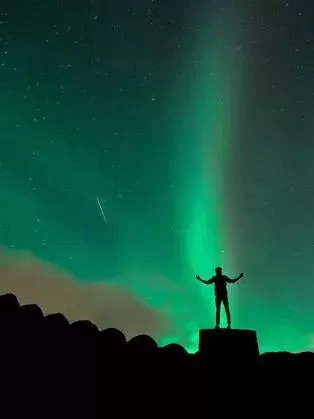 Man Standing With Arms Spread In Joy Underneath the Green Lights of the Aurora Borialis as he watches a shooting star.  Advanced Therapy institute of Touch tries to help others attain their full potential.
